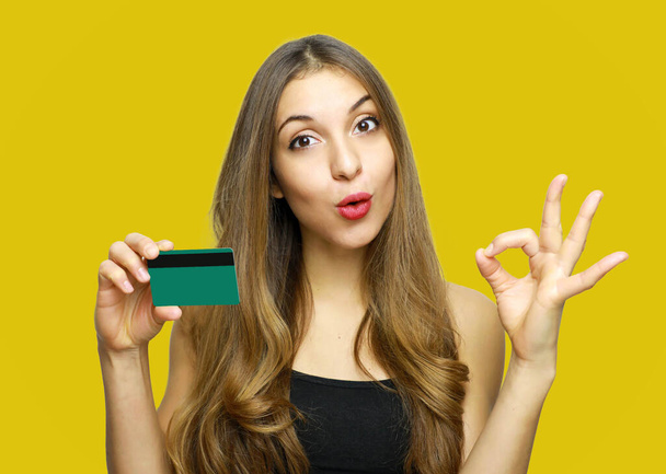 Attractive young woman showing credit card and showing OK sign over yellow background - Foto, Imagen