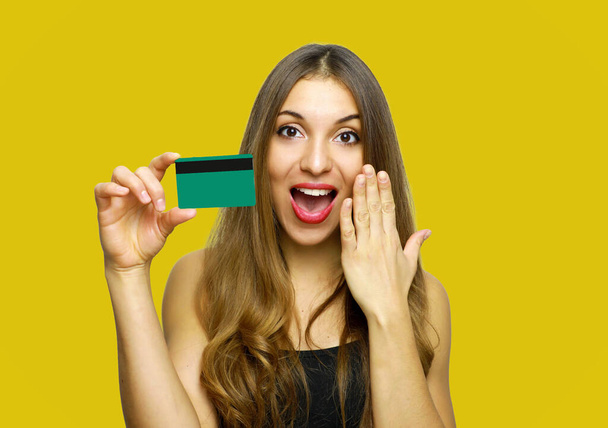 Image of surprised young lady standing over yellow background and holding debit card in hands. Looking at camera. - 写真・画像
