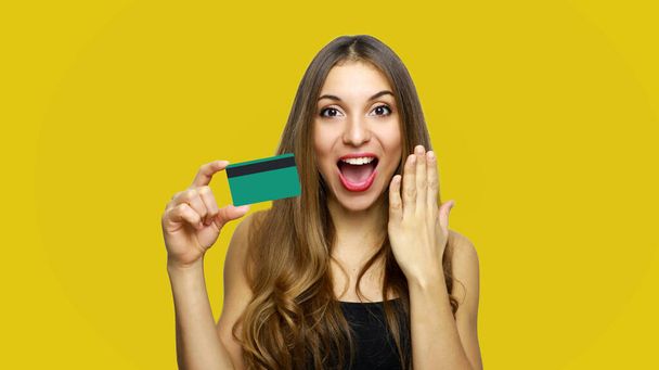 Image of surprised young lady standing over yellow background and holding debit card in hand. Looking at camera with hand near face. - Foto, Imagen