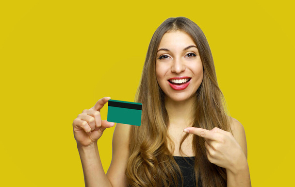 Close up portrait of cheerful young girl with long hair showing and pointing finger credit card isolated over yellow background - Photo, image