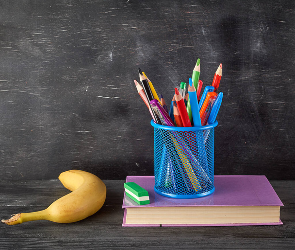 yellow banana, blue stationery glass with multi-colored wooden pencils and pens, on the background of an empty black chalk board, back to school concept - Photo, image