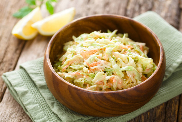 Coleslaw made of freshly shredded white cabbage and grated carrot with homemade mayonnaise-based salad dressing in wooden bowl, lemon wedges in the back (Selective Focus, Focus one third into the salad) - Photo, image