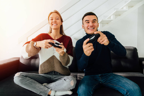 Funny young couple playing computer games with joysticks, sitting on couch, at home. Dressed in sweaters and jeans. - Photo, Image