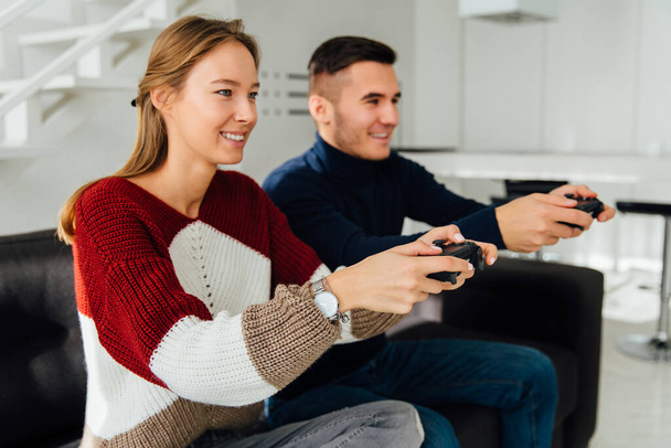 Beautiful young couple playing video games using joysticks, sitting on couch at home. Dressed in warm casual clothes. Side view. - Photo, image