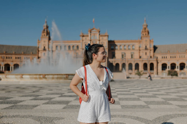 Une jeune touriste portant des lunettes avec une robe blanche et un sac à dos rouge sourit et se couvre les yeux du soleil alors qu'elle se tient debout sur la Plaza de Spain à Séville. Aveuglé par le soleil rude. - Photo, image