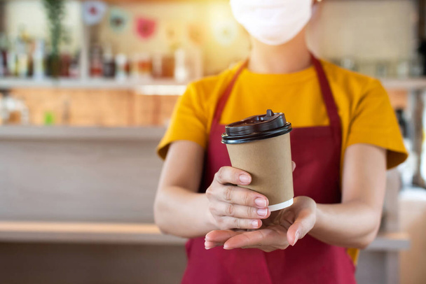 asian woman staff wearing protection face mask waiting for serving hot black coffee cup to customer in cafe coffee shop, cafe restaurant, service mind, new normal, food and drink delivery concept - Photo, Image
