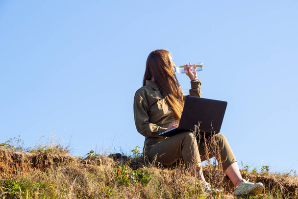 Ein Mädchen mit Laptop sitzt im Gras am Ufer eines Parks außerhalb der Stadt. Das Konzept der Freiberuflichkeit, der Fernarbeit oder der Ausbildung. Eine selbstständige Unternehmerin arbeitet in der Natur auf Reisen. - Foto, Bild