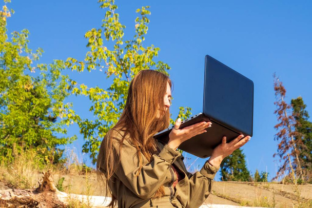 Girl with a laptop is sitting on coastal concrete slabs in a park outside city against background of a forest. Concept of freelance, remote work. Independent female businessman works in nature travel. - Foto, imagen