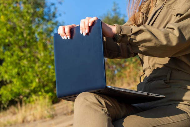 Girl with a laptop is sitting on coastal concrete slabs in a park outside city against background of a forest. Concept of freelance, remote work. Independent female businessman works in nature travel. - Foto, Imagem