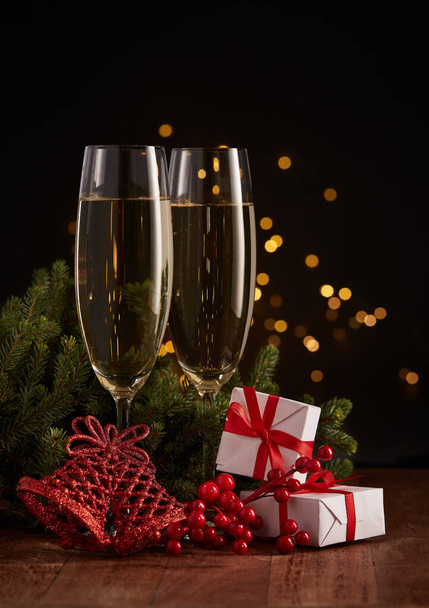 Two glasses with champange, gift boxes, fir tree branch with decorations on a dark background with LED lights garland. New year and Christmas. - Фото, изображение