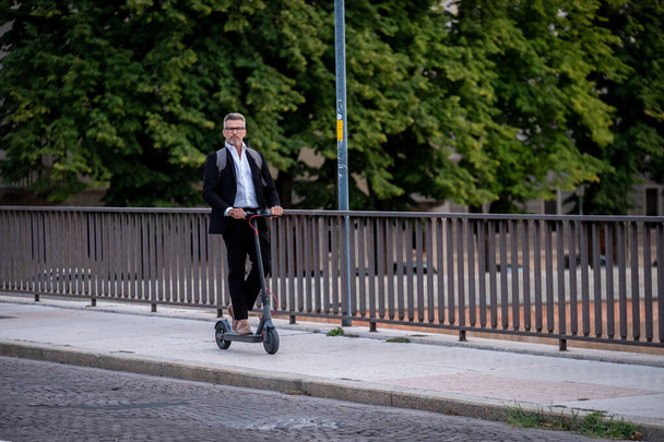 Красивый бизнесмен в костюме ездит на электрическом скутере, когда ездит на работу в город. Концепция экологического транспорта - Фото, изображение