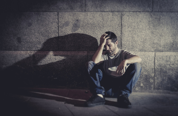 Young man sitting sick on street ground with shadow on concrete wall feeling miserable and sad in urban scene in depression - Foto, Imagem