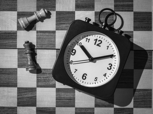 Черно-белое изображение часов на фоне шахматной доски и шахматных фигур. Концепция бизнеса и времени. - Фото, изображение