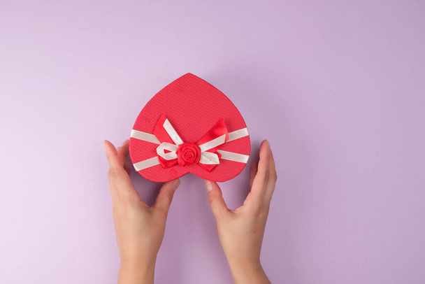 δύο χέρια κρατούν ένα χαρτί κλειστό κόκκινο κουτί σε σχήμα καρδιάς, εορταστική φόντο - Φωτογραφία, εικόνα