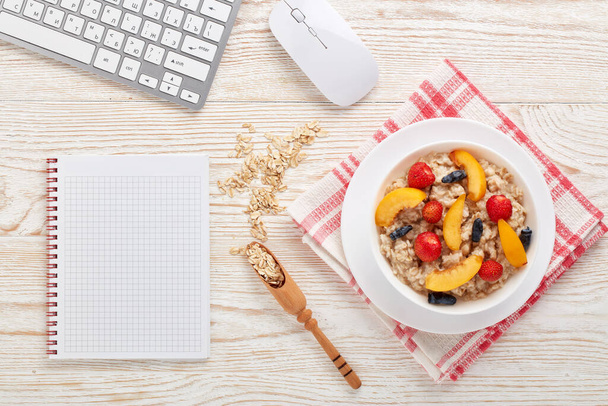 Oatmeal porridge with fresh berries in white bowl,  blank notebook and computer keyboard on light wooden background. Top view. Concept of healthy lifestyle, healthy food, dieting, vegetarian food - Foto, immagini