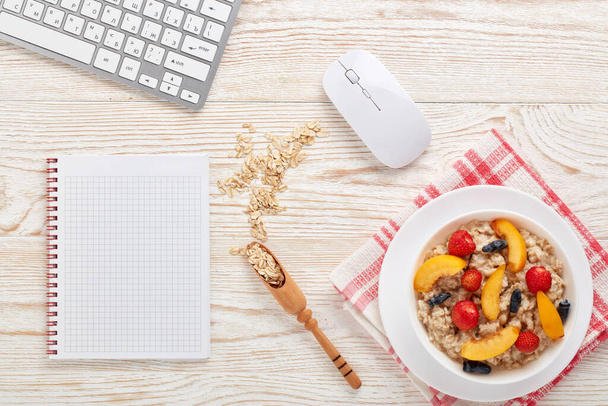 Oatmeal porridge with fresh berries in white bowl,  blank notebook and computer keyboard on light wooden background. Top view. Concept of healthy lifestyle, healthy food, dieting, vegetarian food - Foto, afbeelding