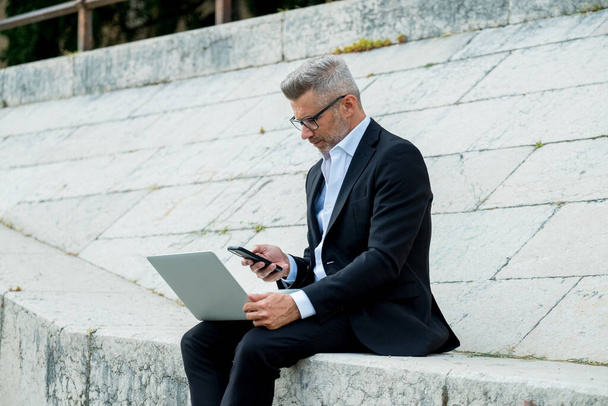 Bello giovane uomo d'affari seduto sulle scale all'aperto che lavora con il suo computer portatile mentre controlla il messaggio al telefono. Lavoro a distanza - Foto, immagini