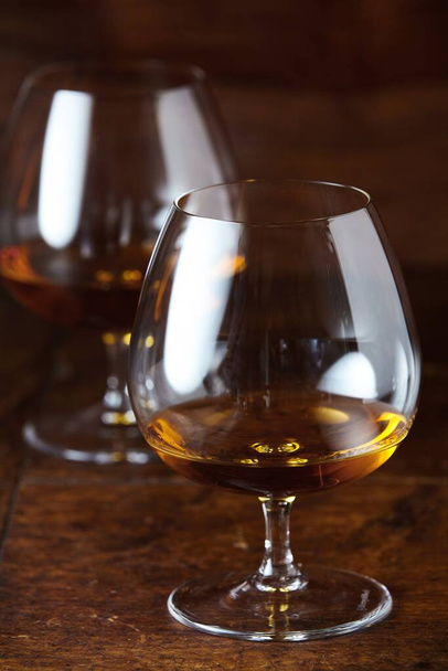 Two filled cognac snifter glasses viewed in close-up on dar brown wooden counter surface in selective focus - 写真・画像
