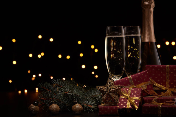 Still life with champagne bottle, two champagne flutes, gift boxes, fir branch, Christmas ornaments and LED lights garland on black background with copy space. Christmas and New Year.  Greeting card - Foto, imagen
