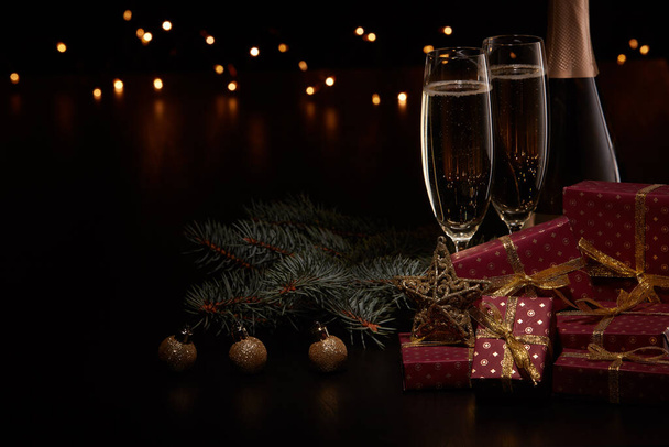 Still life with champagne bottle, two champagne flutes, gift boxes, fir branch, Christmas ornaments and LED lights garland on black background with copy space. Christmas and New Year.  Greeting card - Zdjęcie, obraz