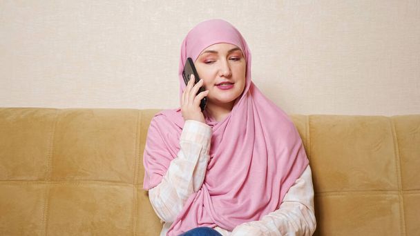 Jovem mulher muçulmana falando ao telefone enquanto se senta no sofá - Foto, Imagem