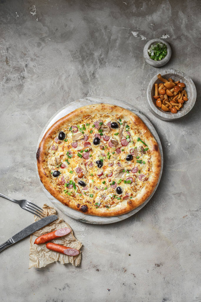 Sosisli lezzetli pizza, mozzarella, marine edilmiş mantar, siyah zeytin, mermer tepside yeşil soğan. Gri taşlı arka planda pizza malzemeleriyle süslenmiş.  - Fotoğraf, Görsel
