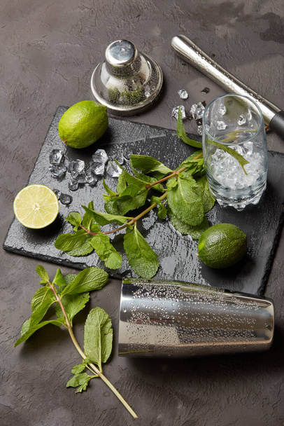 Set of bar tools: cocktail shaker, muddler and glass with ice, lime and mint  on dark stone  table. Top view. Bartender's tool. Mojito cocktail making. - Zdjęcie, obraz