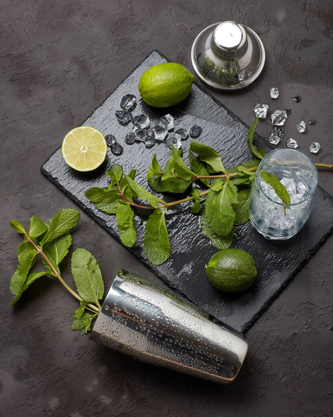 bar tool: cocktail shaker and glass with ice, lime and mint on dark stone  table. Top view. Bartender's tool. Mojito cocktail making. - Fotoğraf, Görsel
