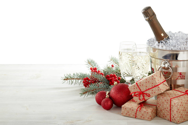 Still life with champagne bottle standing in bucket with ice, champagne flutes, gift boxes, fir branch, Christmas decoration on wooden table on white background with copy space. Christmas and New Year - Photo, image