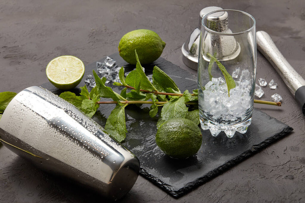 Set of bar tools: cocktail shaker, muddler and glass with ice, lime and mint  on dark stone  table. Top view. Bartender's tool. Mojito cocktail making. - Photo, image