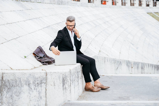 Красивый молодой бизнесмен, сидящий на лестнице и работающий с ноутбуком, разговаривая по телефону. Работа на расстоянии - Фото, изображение