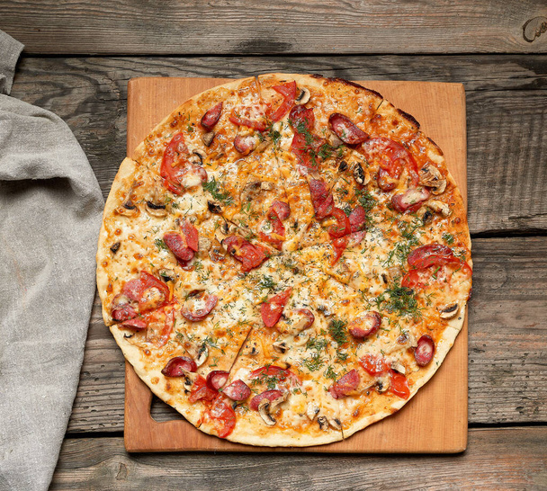 baked round pizza with smoked sausages, mushrooms, tomatoes, cheese, food is cut in portions, wooden table, copy space - Foto, Imagem