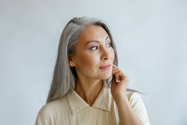 Mujer de pelo gris reflexivo en camisa elegante se ve a un lado en el fondo claro - Foto, imagen