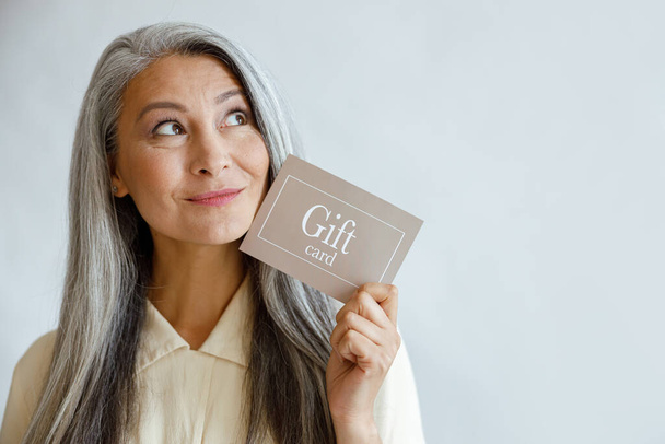 Sonhador de cabelos grisalhos mulher asiática detém cartão de presente perto do rosto no estúdio - Foto, Imagem