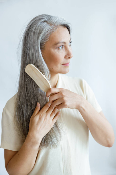 Languid middle aged Asian woman brushes natural hoary hair on light grey background - Foto, Imagen
