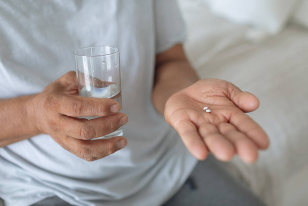 Close up of senior Caucasian man sitting on the bed while holding pills and a glass of water. Authentic Senior Retired Life Concept - Photo, Image