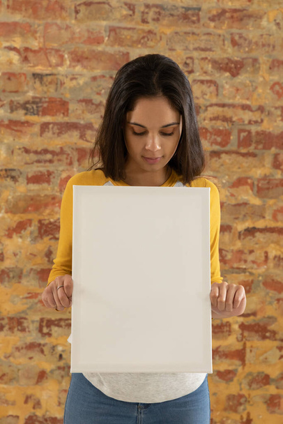 Front view close up of a young Caucasian woman holding a blank white canvas in her hands and looking down at it, standing in front of a brick wall - Photo, image