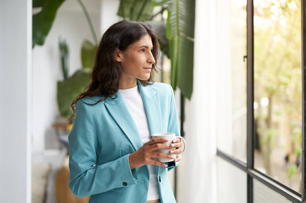 Young business woman satisfied with a job well done relaxing with her morning coffee or tea, looking out the window. Beautiful latina woman celebrating successes on break from work at the office. - Photo, Image