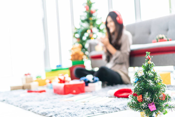 Here is a picture of a small Christmas tree in a white room. The backdrop of the Christmas tree depicts a woman sitting listening to music with a red headphones and having many gift boxes. - Foto, immagini
