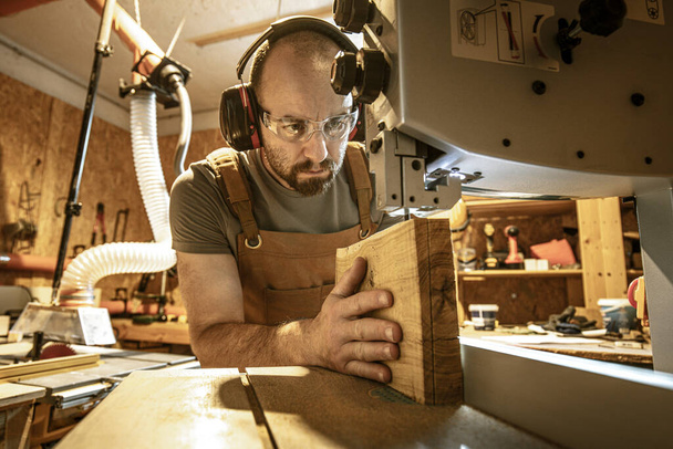 portrait of a carpenter inside his carpentry workshop using a band saw. Wear ear and eye protection - 写真・画像
