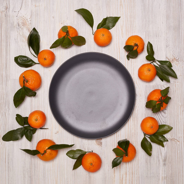 Frame from ripe mandarins with leaves and black empty plate on light wooden background. Top view with copy space. Festive background from tangerines - Foto, imagen