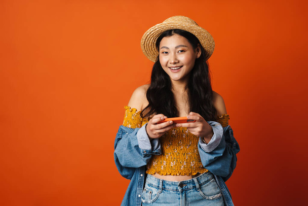 Portrait of a happy young brunette asian woman in summer hat standing over orange wall background, holding mobile phone - Photo, Image