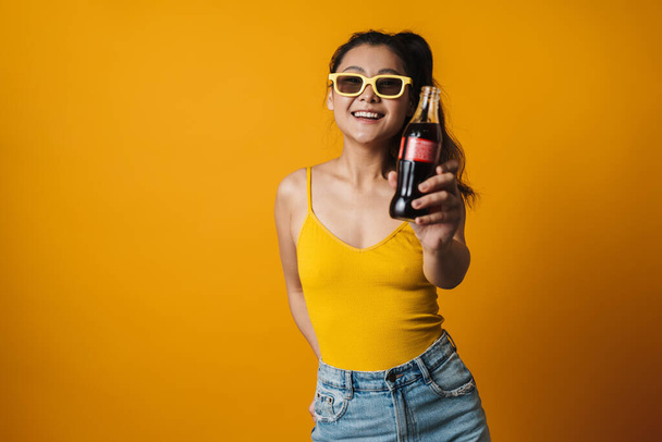 Happy young asian woman wearing summer clothes over orange background, holding bottle with a fizzy drink - Foto, imagen