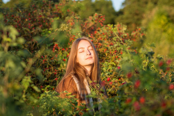 Ein helles, warmes Herbstporträt eines jungen kaukasischen Mädchens in einem ledernen Schaffellmantel vor einem Hintergrund aus roten Sträuchern und Hagebuttenbeeren. Eine Frau sonnt sich in den Strahlen der untergehenden Sonne. - Foto, Bild