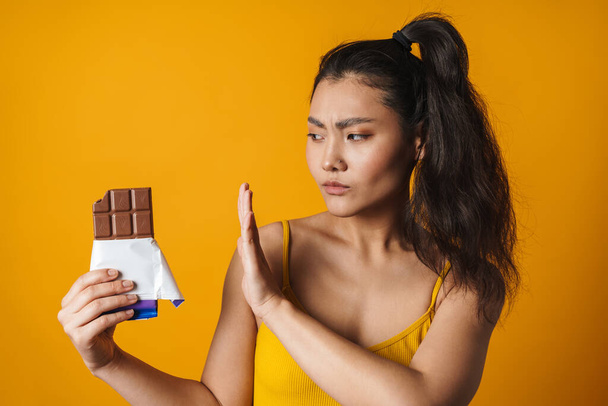 Portrait of an upset young asian woman holding chocolate bar standing over yellow background - Фото, зображення