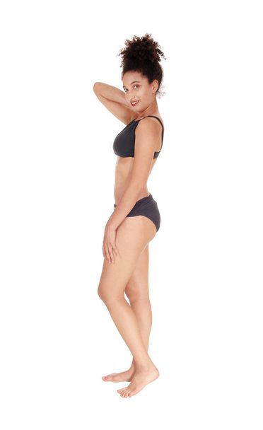 A beautiful slim young multi-racial woman standing in profile in a black.bikini in the studio looking into camera, bare foot with her curly black .hair in a bun on her head, isolated for white background. - Foto, Imagen