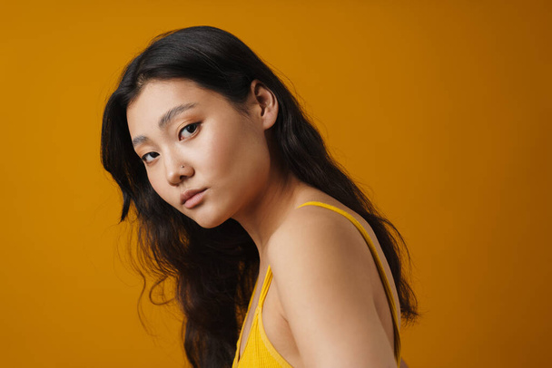 Portrait of a young brunette asian woman in top posing standing over yellow background - Fotografie, Obrázek