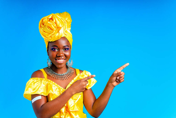 beautiful african woman in yellow silk turban on head and dress with silver neckless and earrings , nose piercing in blue studio background - Fotografie, Obrázek