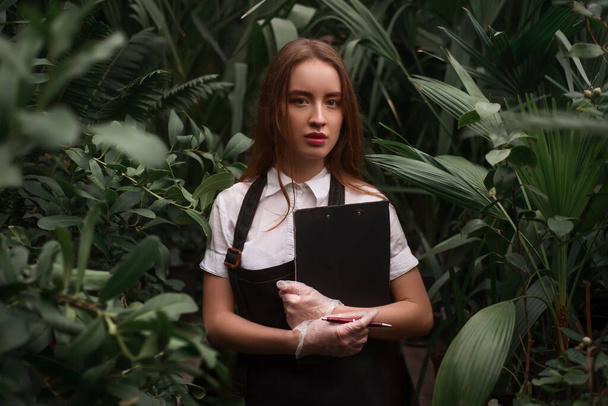 Young florist in apron holds clipboard in hands against greenhouse with exotic plants. - Foto, immagini