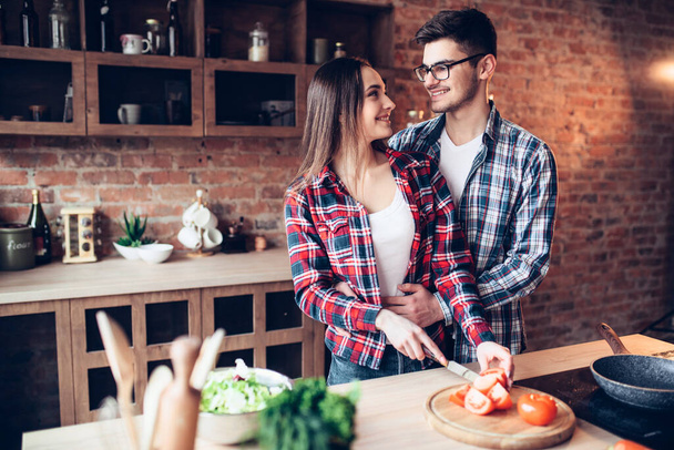 Husband hugs wife while she cooking fresh vegetable salad in bowl on the kitchen. Diet food preparation. Couple prepares romantic dinner, healthy lifestyle - Fotó, kép